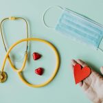 Top view of crop anonymous person hand with red paper heart on table with stethoscope and medical mask for coronavirus prevention